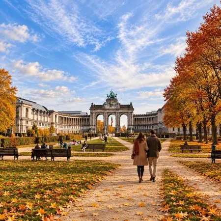 Couple walking down a tree-lined autumn park toward a grand arched monument across a wide plaza.